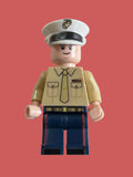 A United States Marine toy minifigure shown from the side on a plain light background. The figure has a beige upper torso with printed pockets and tie details, dark blue lower body panels, a white hat, and pale peach skin.
