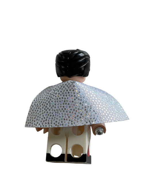 A miniature toy figure viewed from the back, featuring a black hairpiece, a silver patterned cape, and a cream-colored torso with circular cutouts, set against a plain white background.