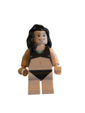 A small toy minifigure with a beige torso and legs, dark brown hair, and a black-and-beige outfit, photographed on a plain light background.