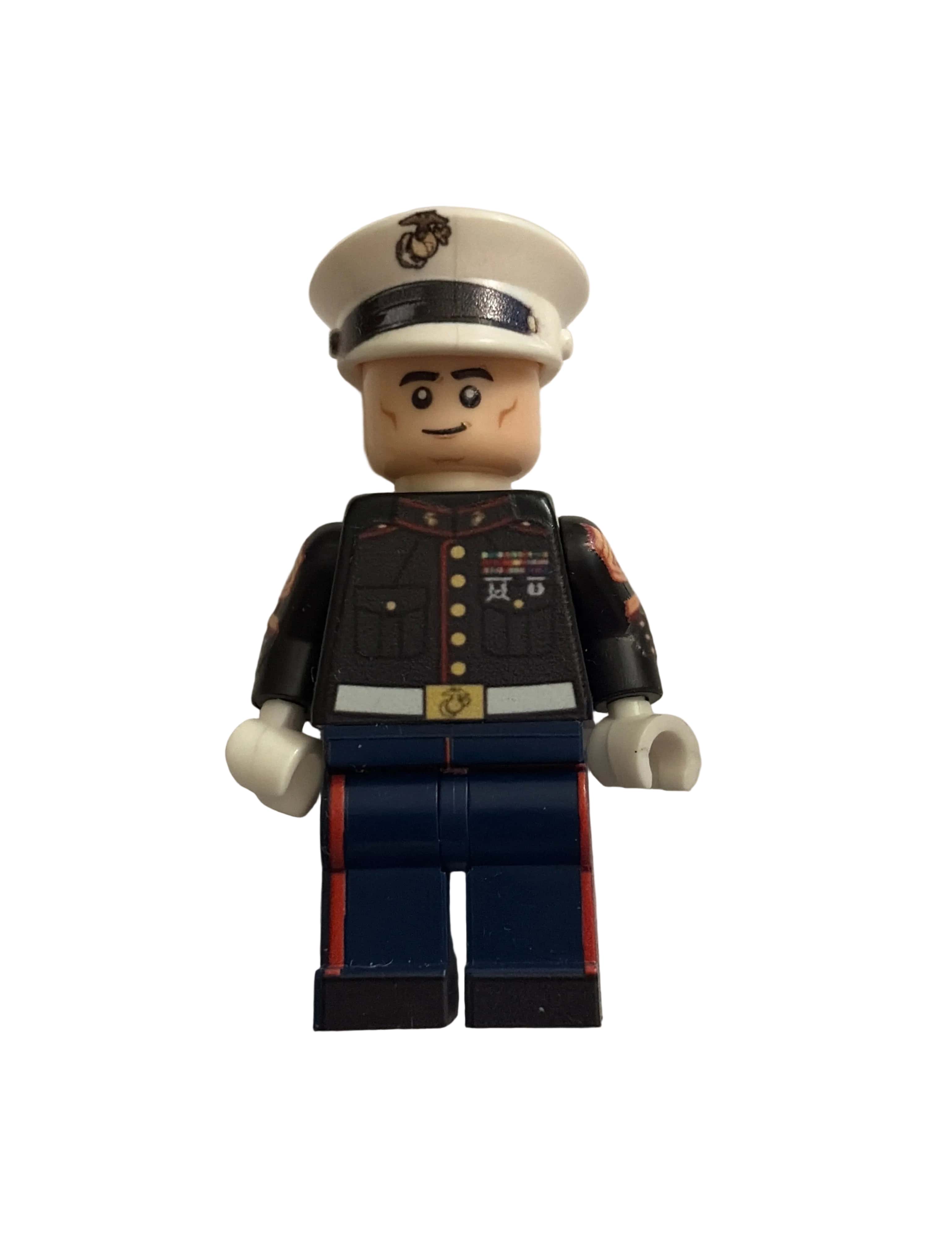 A toy United States Marine military minifigure with a beige head and hands, wearing a white service hat, a black jacket with gold buttons, and dark blue pants with red stripes, isolated on a white background.