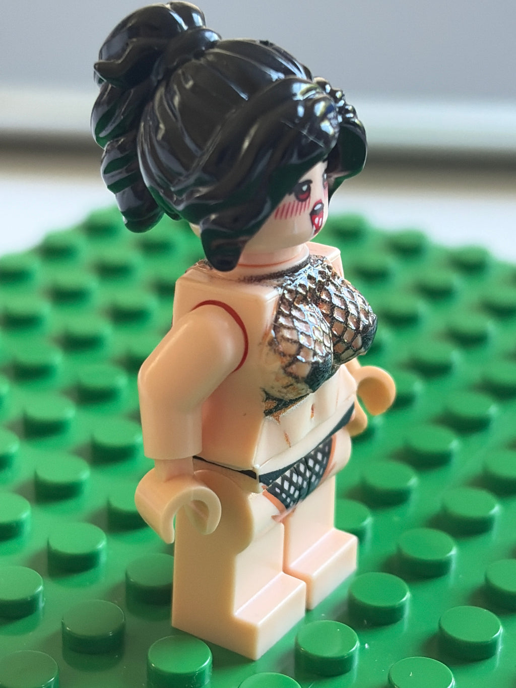 A custom-built female LEGO minifigure with black hair in a high ponytail, wearing a fish-scale patterned bikini top and black patterned bikini bottom, standing on a green LEGO base plate.
