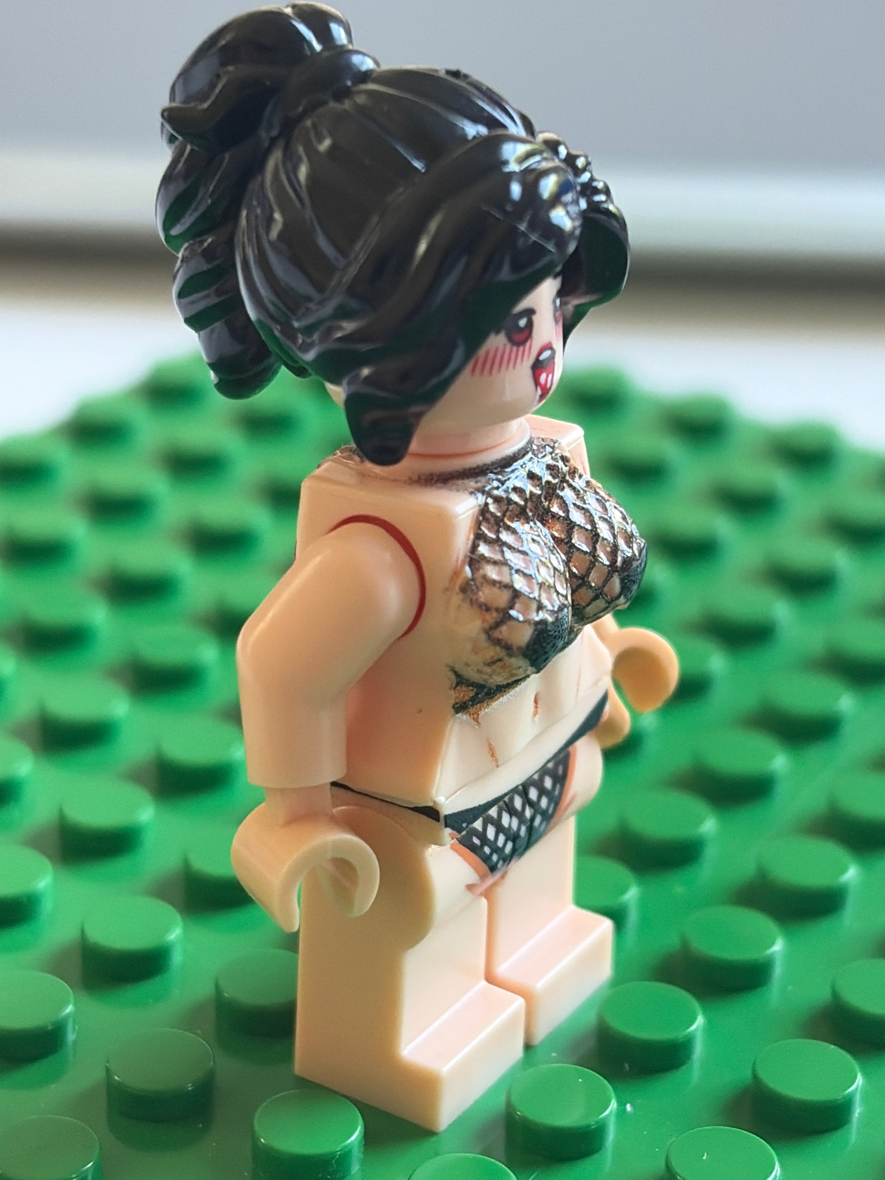 A custom-built female LEGO minifigure with black hair in a high ponytail, wearing a fish-scale patterned bikini top and black patterned bikini bottom, standing on a green LEGO base plate.