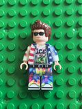 A custom-built toy minifigure with brown hair, black sunglasses, a red, white, and blue star-patterned jacket, a graphic t-shirt, and colorful holographic pants, standing on a green LEGO-style baseplate.