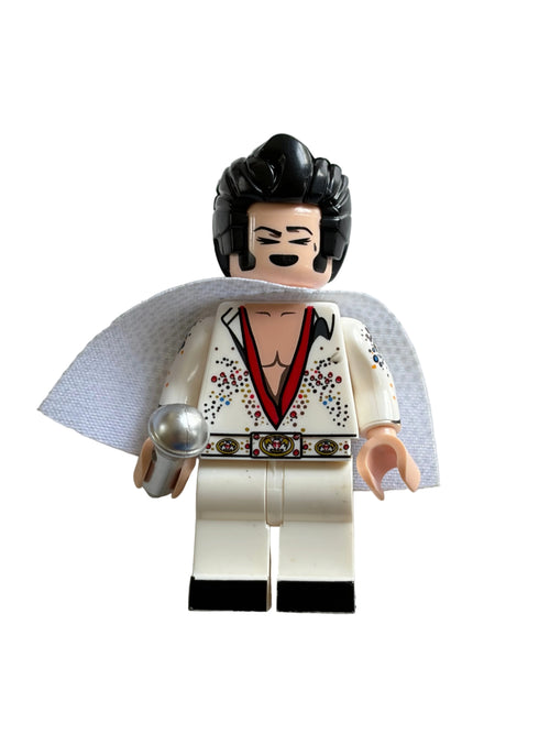 A custom-built mini figure of Elvis Presley, styled  minifigure, wearing a white jumpsuit with red and black embroidery, a white cape, and black hair, holding a silver microphone against a white background.