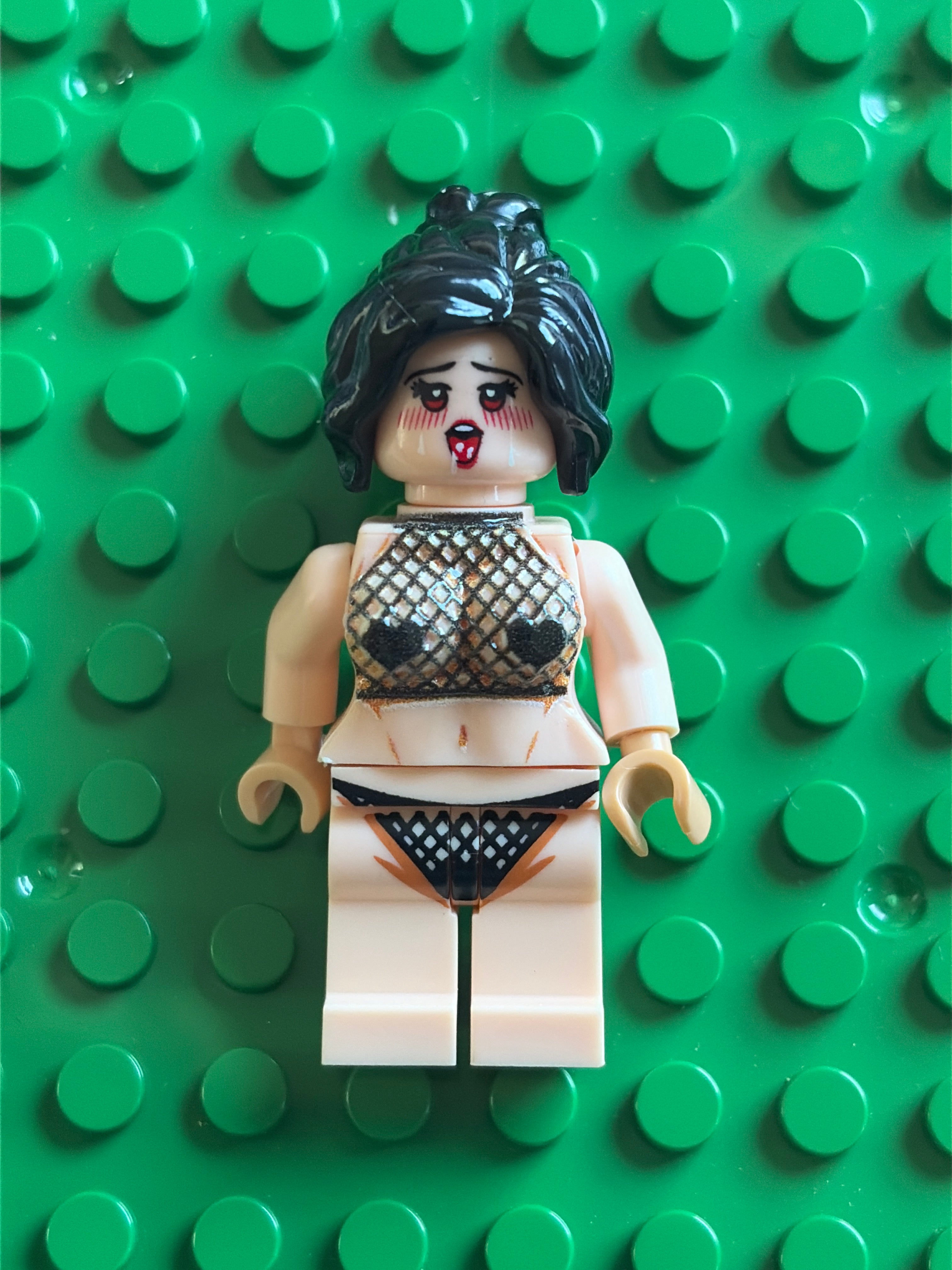 A custom-built toy minifigure with a distressed beige skin tone, black hair, and a bloody black-and-white patterned bikini, standing on a green LEGO brick baseplate.