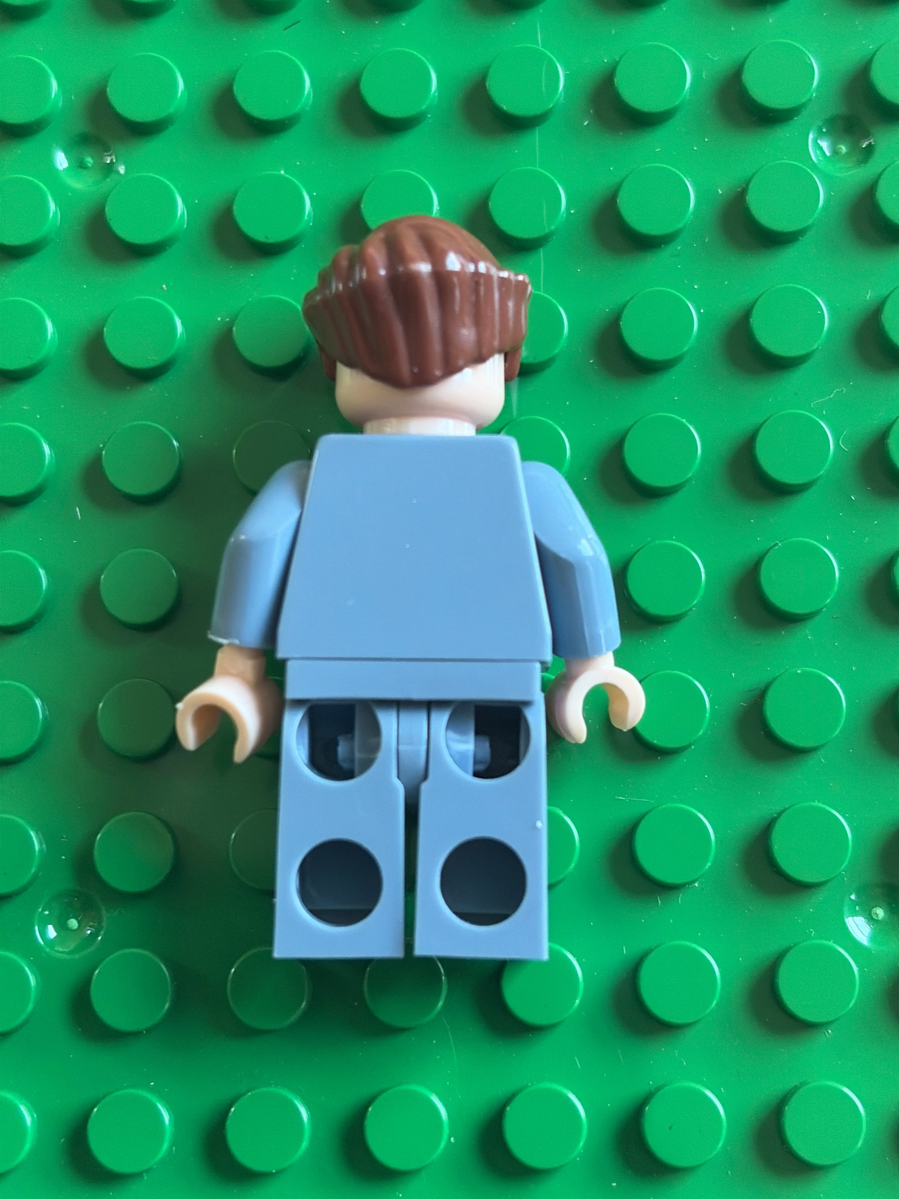A small toy minifigure viewed from the back, with brown hair, a light blue torso and legs, and beige hands, placed on a bright green stud base plate.