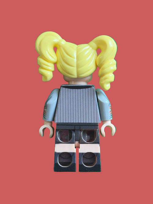 A custom LEGO minifigure viewed from the back, featuring bright yellow pigtails, a gray ribbed top, light blue sleeves, and black shorts, standing on a green LEGO brick base.