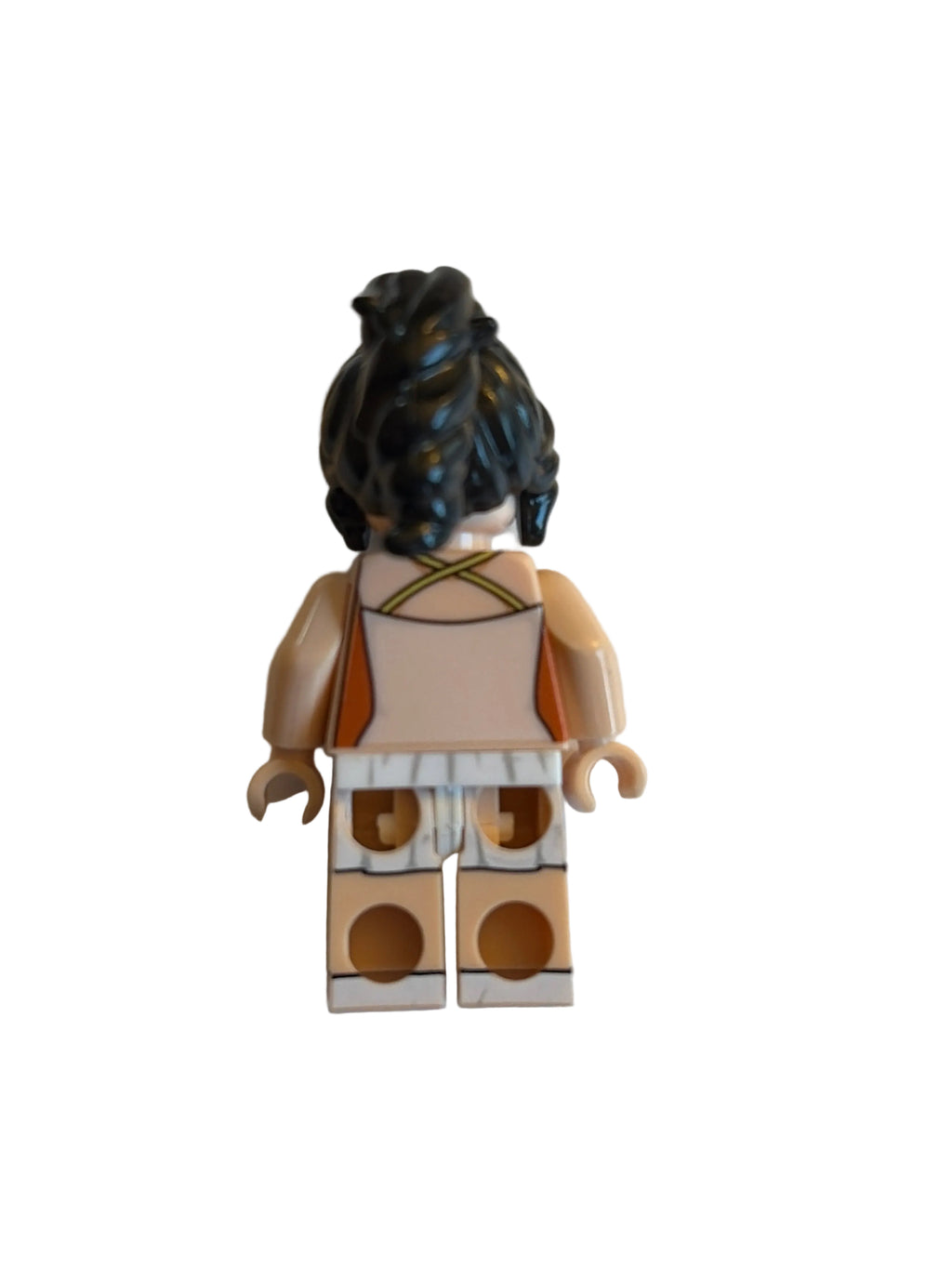 A small toy minifigure shown from the back on a plain off-white background. The figure has dark curly hair, a tan and white outfit with brown accents, and beige plastic limbs and torso.