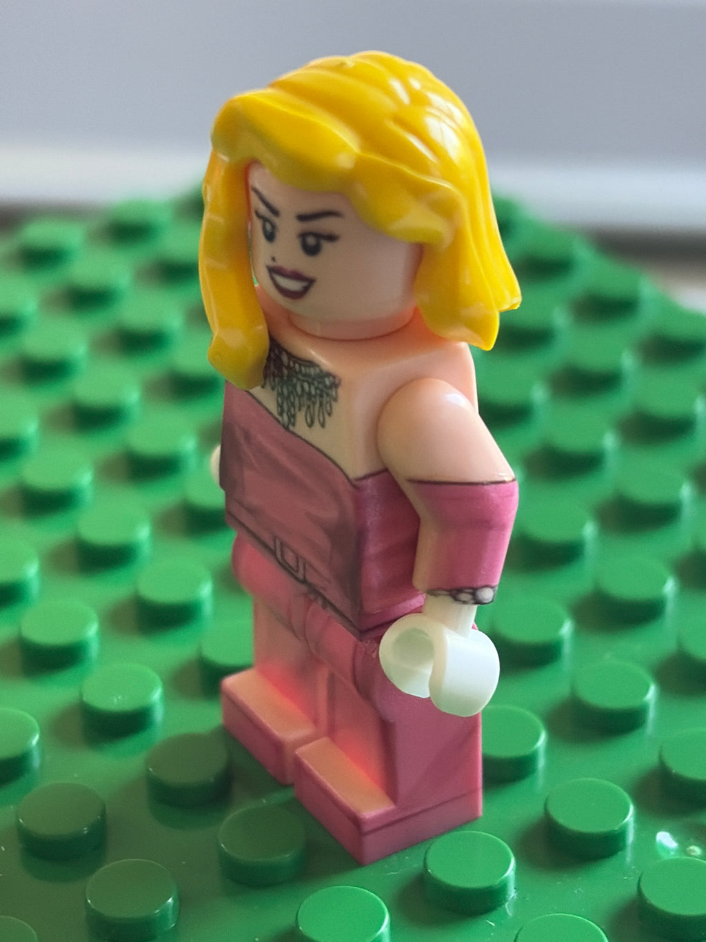 A custom-built LEGO minifigure with blonde hair, a pink top, and pink pants, standing on a green LEGO baseplate.