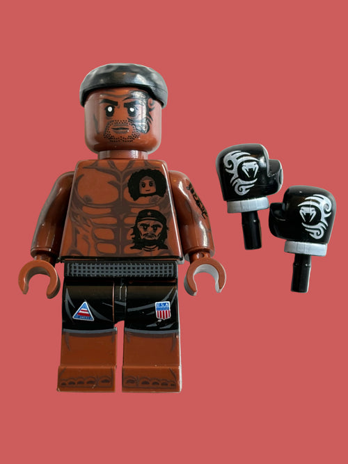 A custom-built brown brick minifigure with a black cap, a printed muscular torso, and black pants, shown alongside a pair of black and white printed boxing gloves on a light beige background.