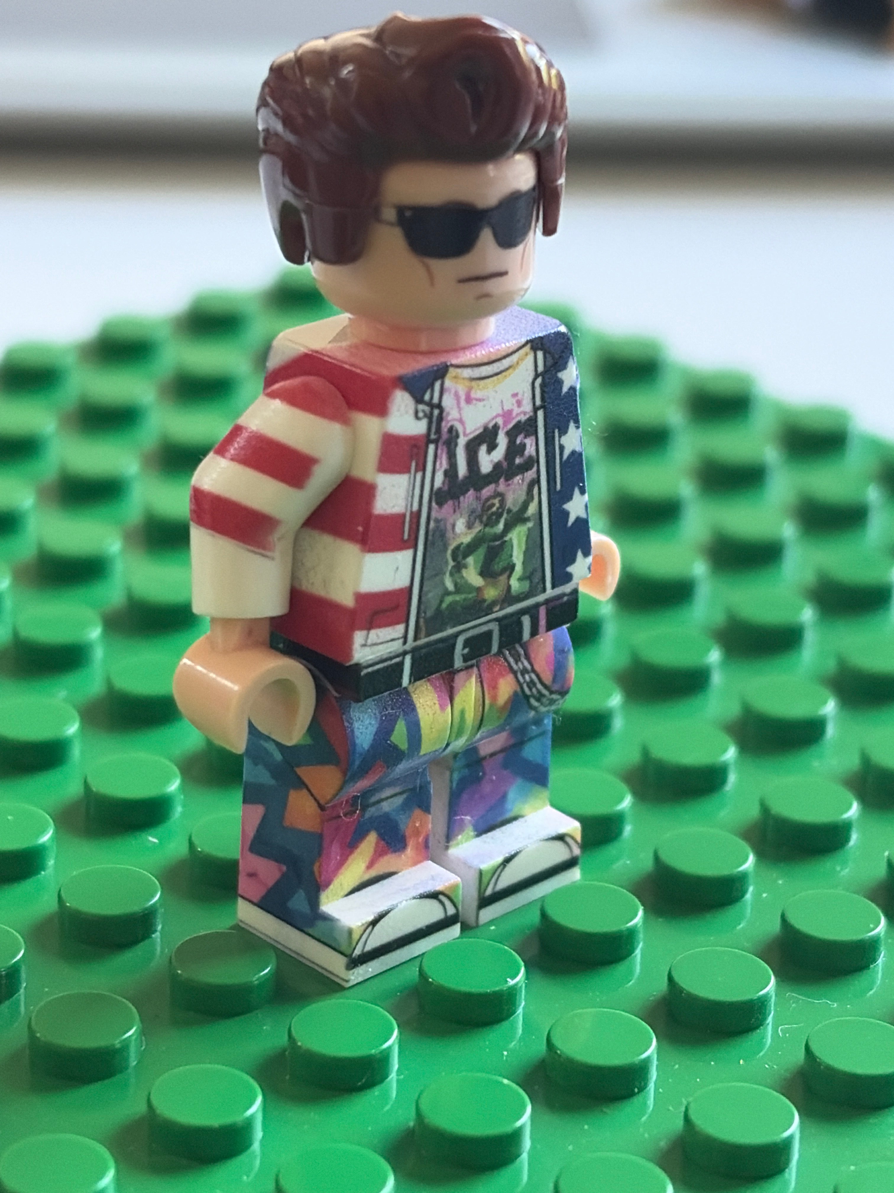 A custom-built toy minifigure with brown hair, black sunglasses, a red and white striped shirt, and a colorful graphic jacket and patterned pants, standing on a green LEGO baseplate.