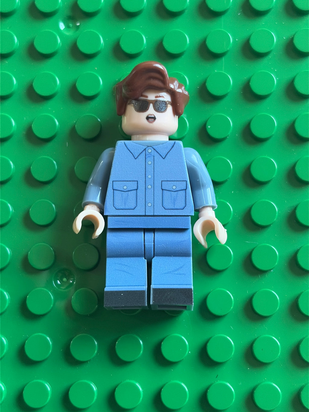 A custom LEGO minifigure with brown hair, sunglasses, and a blue jumpsuit, standing on a green LEGO baseplate.