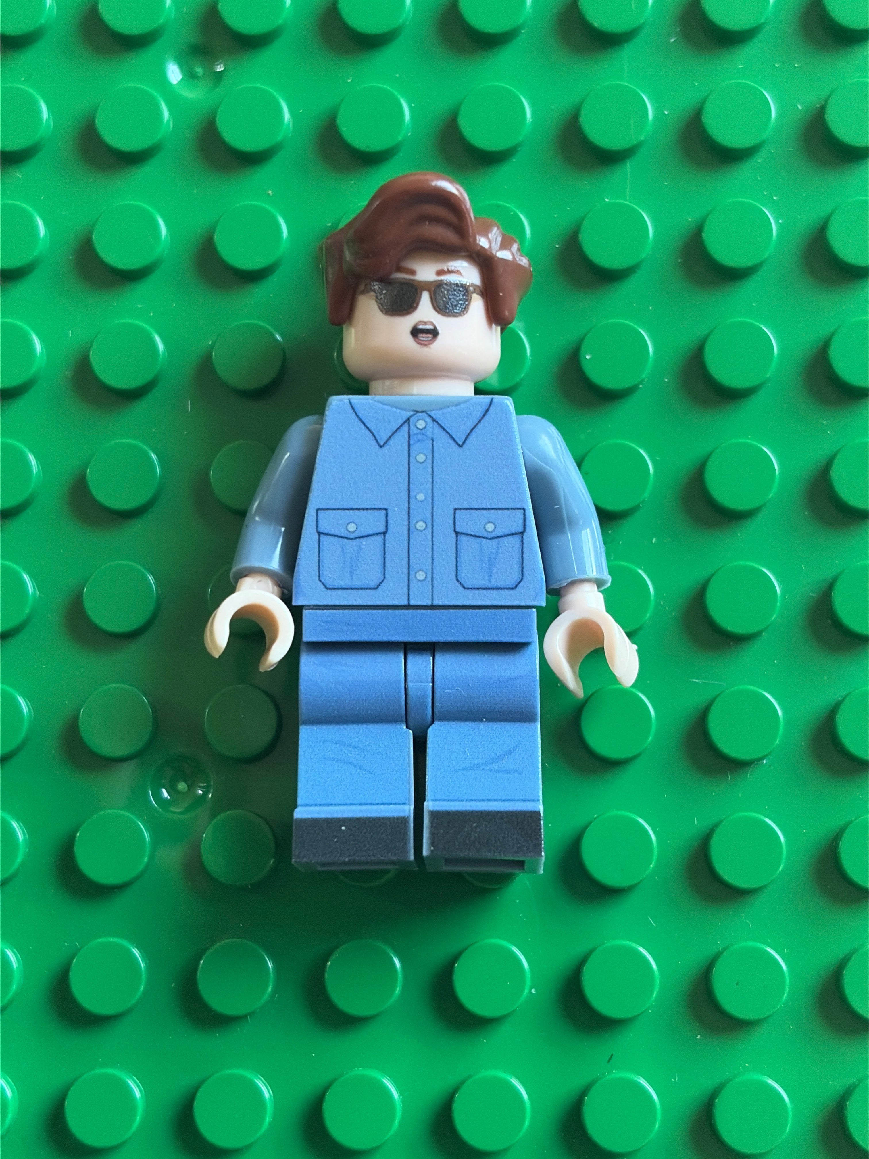 A custom LEGO minifigure with brown hair, sunglasses, and a blue jumpsuit, standing on a green LEGO baseplate.