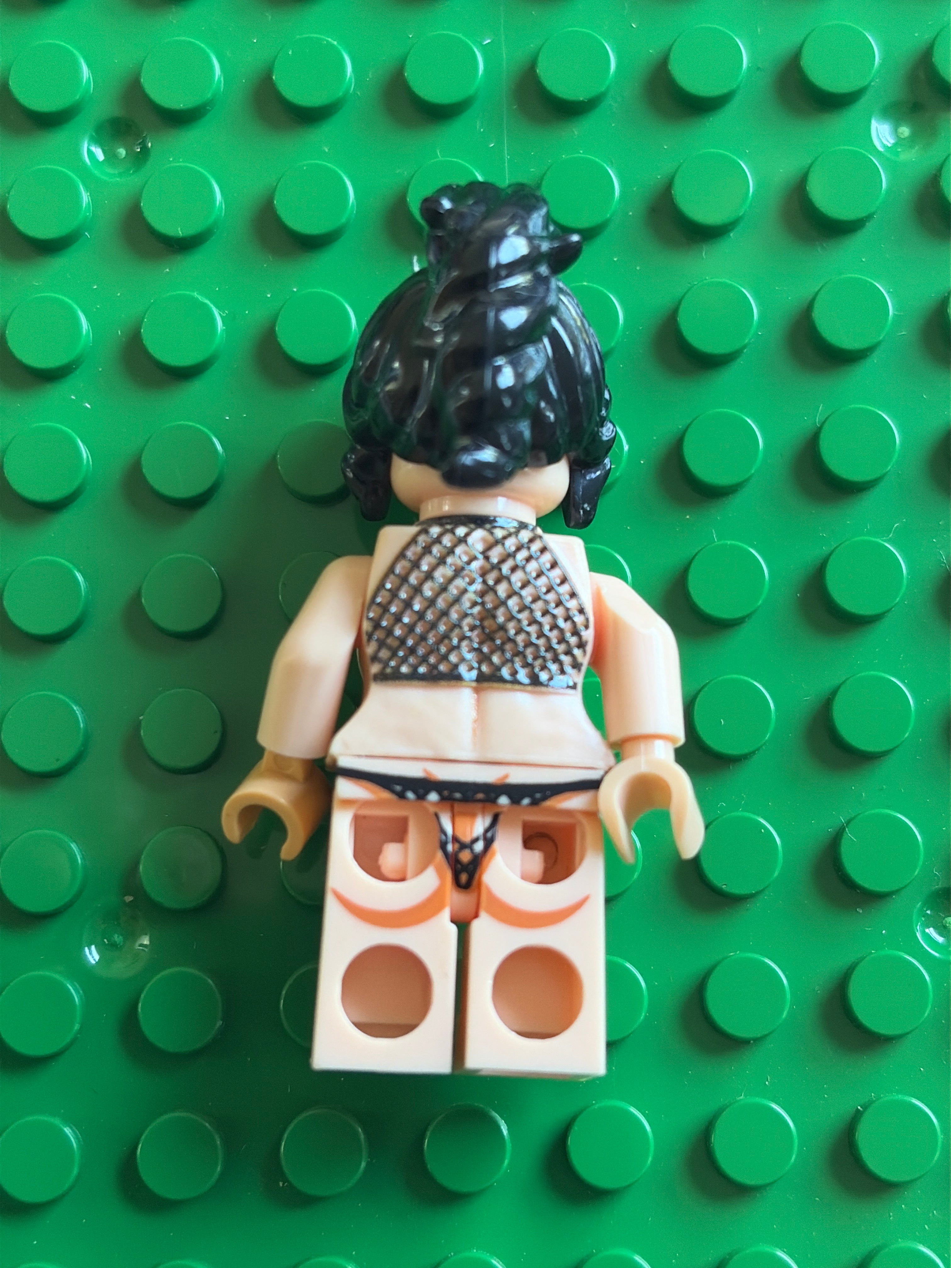 A LEGO minifigure viewed from the back, wearing a black mesh top and black underwear, standing on a green LEGO brick baseplate.