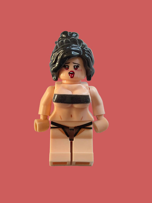 A custom-built toy minifigure with a beige plastic torso and legs, black hair, and a black and beige lingerie set, lying on a light pink surface.