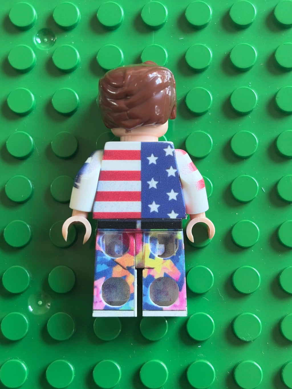 A custom LEGO minifigure viewed from the back, featuring brown hair, a white and blue American flag-themed torso, and colorful tie-dye patterned legs, set against a green LEGO brick base.