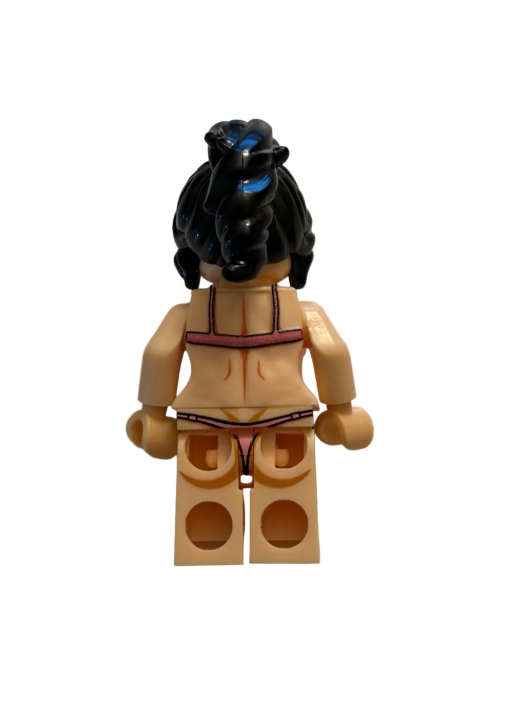 A small toy figure or accessory piece lying on a plain light gray surface, with a black hair section, beige body parts, and a tan rectangular block piece with circular cutouts.