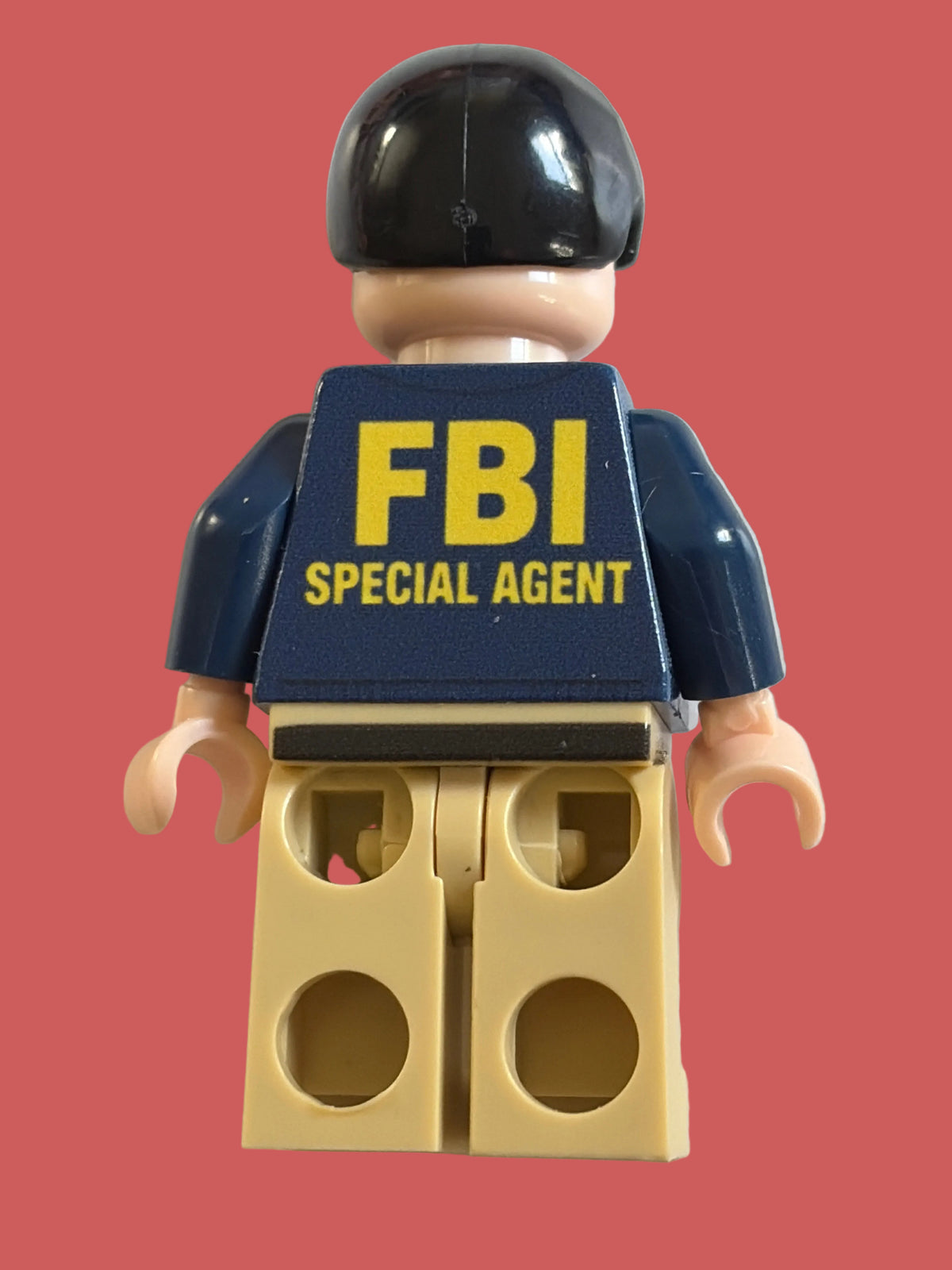 A toy minifigure shown from the back, wearing a black helmet, navy blue shirt with yellow 'FBI SPECIAL AGENT' text, and tan pants, photographed against a plain light background.