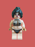 A custom-built toy minifigure with a distressed beige skin tone, black hair, and a mesh black-and-white patterned bikini on a pink background.