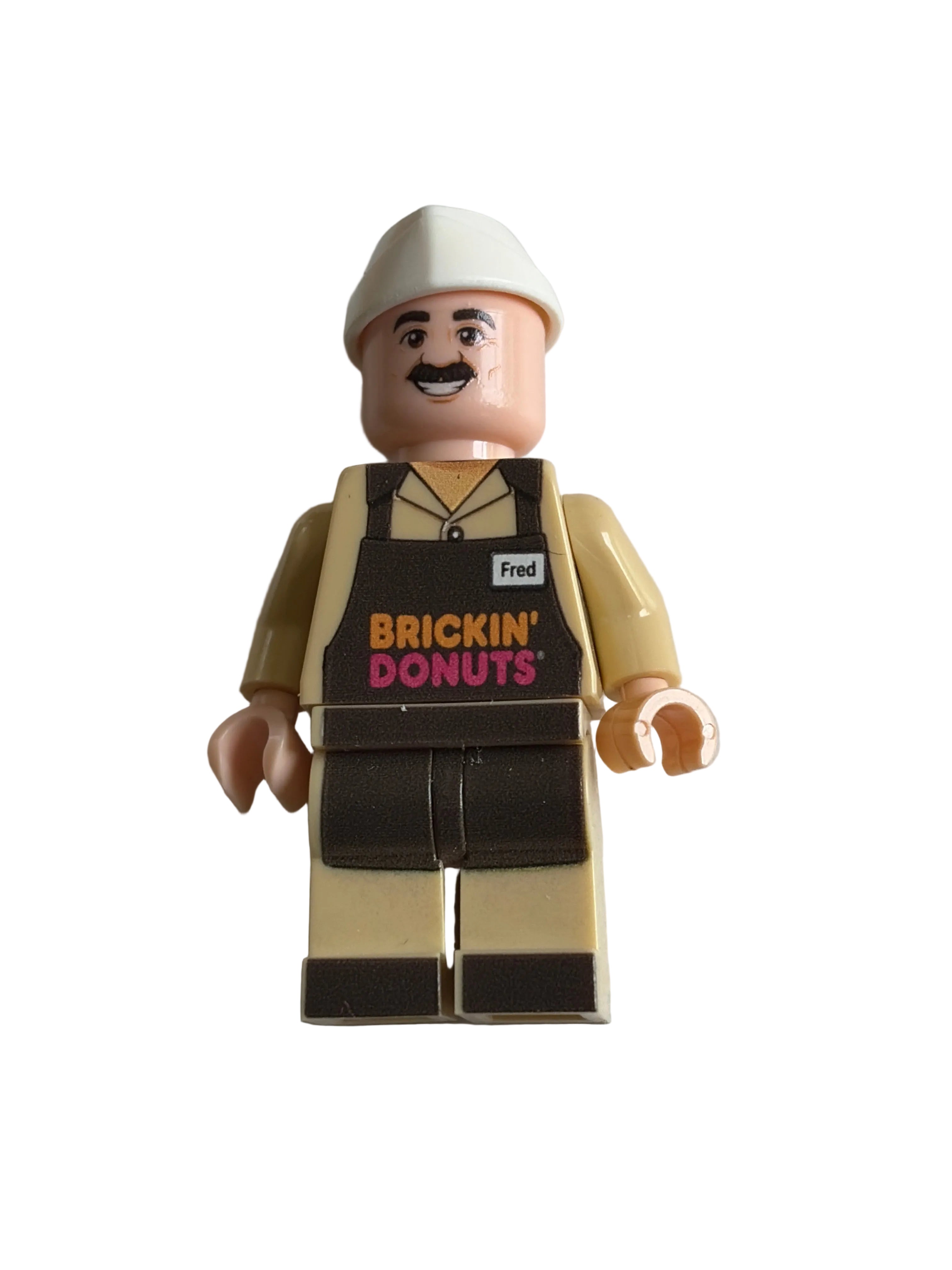 A minifigire of the iconic tv ad character from Dunkin Donut as a parody Brickin Donuts with a white wedge hat, peach skin-tone arms and legs, and a beige outfit and brown apron, photographed on a plain white background.