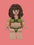 Custom Lego style minifigure peach color wearing a green strapless bikini top and green bikini bottoms, with brown wavy hair, on a pink backgrojnd