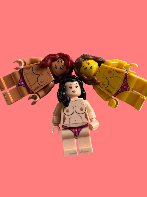 Three toy minifigures posed together on a light surface, wearing purple bikini-style bottoms. Two figures are reclining, one standing upright in front, with varied skin tones and hair colors.