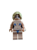 A toy minifigure with blonde hair, blue eyes, and a light flesh skin tone, wearing a blue bikini, lying on a white surface.